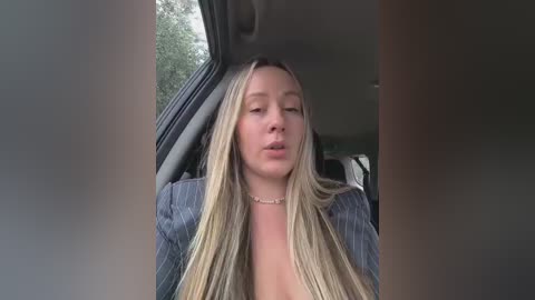 A video of a blonde woman with long hair, wearing a blue pinstripe blazer, seated in a car, looking straight ahead with a neutral expression.