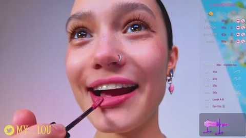 A video of a young woman with fair skin, brown eyes, and dark hair in a ponytail, smiling while applying red lipstick. She wears a nose ring and small earrings. Background includes a map and a digital display.