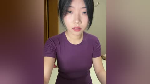 Video of an East Asian woman with fair skin, black hair, and red lips, wearing a tight, purple t-shirt, sitting indoors with blurred background.