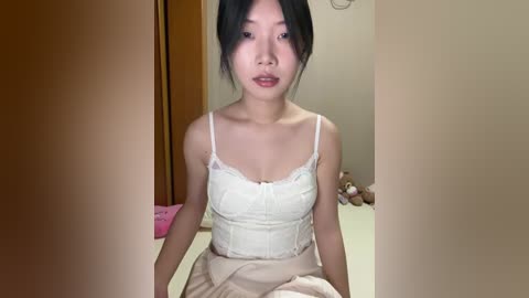 Video of an Asian woman with black hair, wearing a white lace camisole, sitting on a bed with beige sheets, in a dimly lit room with wooden furniture and stuffed animals.