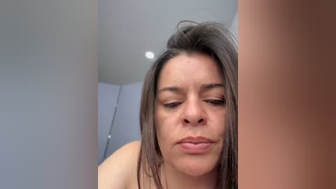 A close-up video of a woman with medium brown skin and straight, shoulder-length dark brown hair, slightly tousled. She has a neutral expression, possibly smiling slightly. The background shows a beige wall and a white ceiling.