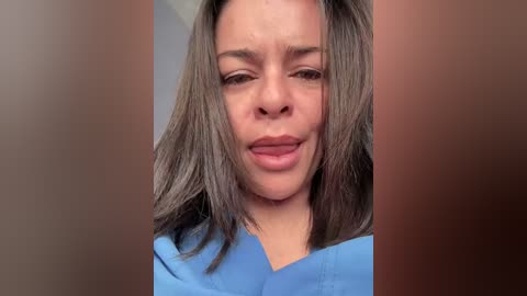 Video of a light-skinned woman with shoulder-length, straight brown hair, wearing blue scrubs, smiling and sticking her tongue out, against a blurred background.