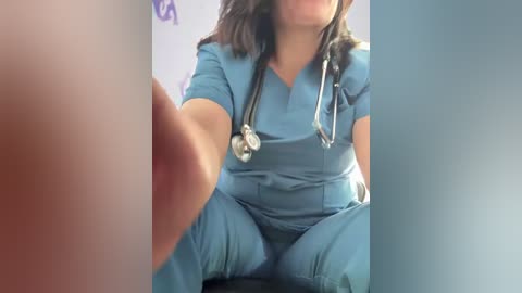 Video of a light-skinned woman with medium-length dark hair, wearing blue scrubs, sitting with legs spread. She has a stethoscope around her neck. The background is blurred.