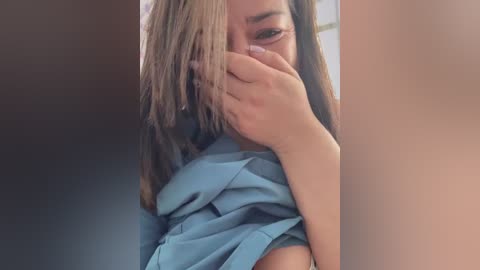 Video of a young woman with light brown skin and shoulder-length, straight brown hair, partially concealing her face with her hand. She wears a light blue shirt, with the background consisting of muted brown and beige tones.