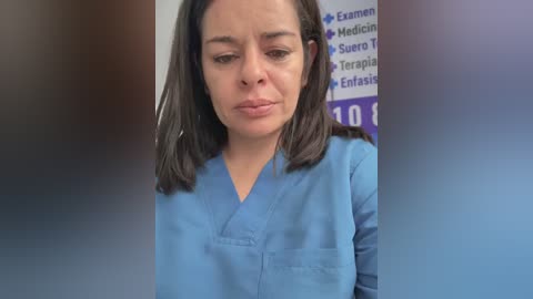 Video of a woman with medium skin tone, dark hair, wearing blue scrubs, looking down, in a medical setting with a blurred background featuring medical terms.