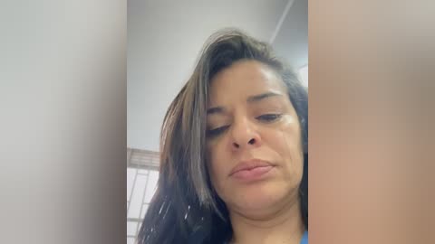 A close-up video of a Latina woman with medium-brown skin, long dark hair, and closed eyes, captured in a dimly lit room with white walls and a window.