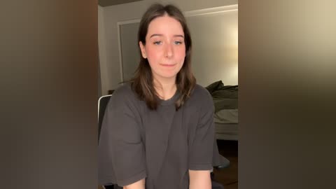 Video of a young Caucasian woman with shoulder-length brown hair, wearing a gray T-shirt, sitting indoors with a blurred background.