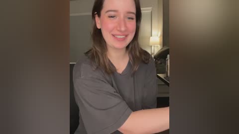 Video of a young woman with shoulder-length brown hair, fair skin, and a casual grey t-shirt, smiling slightly, indoors with a warm, dimly lit background featuring a lamp and wall.