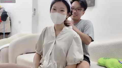 A video of an Asian woman in a white mask and robe being combed by an Asian man in glasses, sitting on a cream sofa in a minimalist living room.