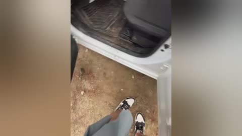 A video of a person's feet, wearing black and white sneakers, standing near the open door of a white car. The ground is dusty, with scattered debris. The image is slightly blurred.