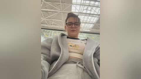 Video of a young woman with glasses and a grey hoodie, sitting in a modern, white, high-ceilinged room with large glass windows.