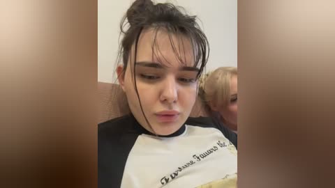 A video of a young woman with dark hair in a messy bun, wearing a black and white shirt, looking downcast, seated on a plane.