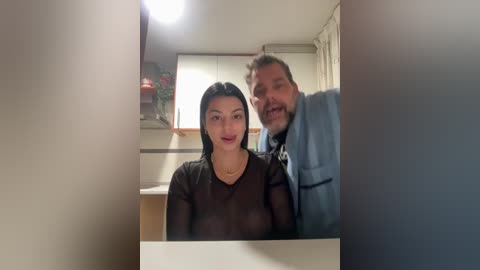 Video of a smiling Asian woman with long black hair and medium breasts wearing a sheer black top, seated in a modern kitchen, with a bearded man in a blue shirt standing behind her.