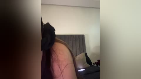 Video of a person with a visible tattoo on their arm, lying on a bed with a grey headboard. The room is dimly lit with a beige wall and a black pillow in the background.