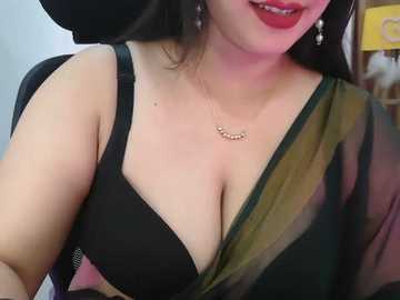 Video of a smiling, fair-skinned woman with dark hair, wearing a black bra and green and black sheer blouse, with dangling earrings.