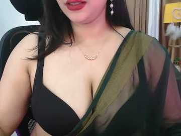 A video of a woman with fair skin and long black hair, wearing a black bra and a semi-transparent green and gold sari, accessorized with a delicate necklace.
