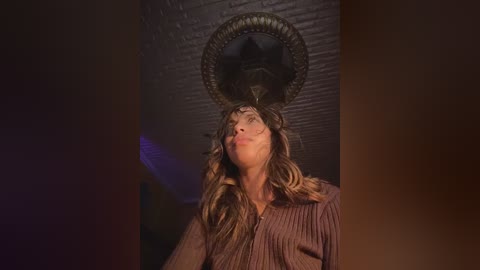 Video of a woman with long, wavy blonde hair, wearing a brown striped shirt, standing in a dimly lit room with a shadowy, ornate ceiling fixture.