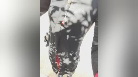 Video of a person wearing a black and white patterned jacket with red accents, standing in a sunlit outdoor area with dappled light.