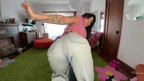 Video of a curvy woman with tattoos, wearing a pink top and light-wash jeans, posing in a colorful living room with a green carpet, red bean bag, and floral decor.