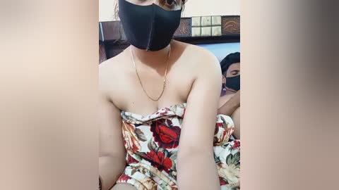 Video of a woman with light brown skin, wearing a black face mask, floral-patterned blanket, and gold necklace, lying on a bed in a room with a wooden headboard and a masked person in the background.