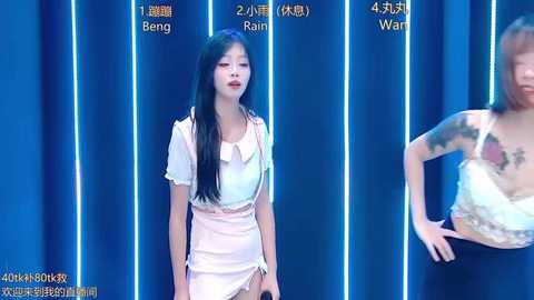 Video of an Asian woman with long black hair and fair skin, wearing a white dress, standing in front of blue curtains.