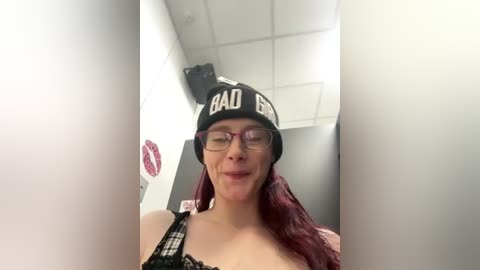 Video of a smiling woman with fair skin and long red hair, wearing glasses, a black \"BAD BLOOD\" beanie, and a black lace top. She stands in a fluorescent-lit room with white walls and a ceiling.