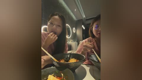 Video of two young Asian women eating noodles in a modern, dimly lit restaurant with round, glowing light fixtures on the wall.