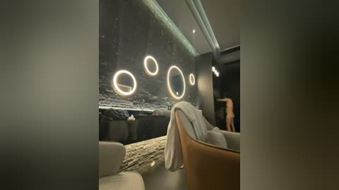 Video of a modern, minimalist room with a snow-covered outdoor view through circular glass windows. Inside, a beige sofa with a white blanket and a wooden armchair are arranged near a wall of glowing, circular light fixtures. The room has a sleek, contemporary design.