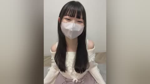A video of an East Asian woman with long, straight black hair and pale skin, wearing a white off-shoulder sweater, face mask, and a pink dress. She stands in a plain, light-colored room with a beige floor.