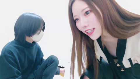 A video of a woman with long brown hair in a black dress, smiling beside a man in a black panda suit, seated on a white floor.