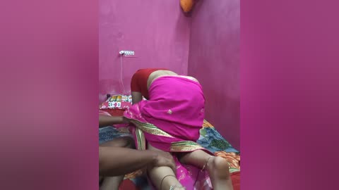 A video of a woman in a bright pink sari, bent over, being held up by two men, in a small, pink-walled room with a colorful bedspread and a partially visible electrical device on the wall.