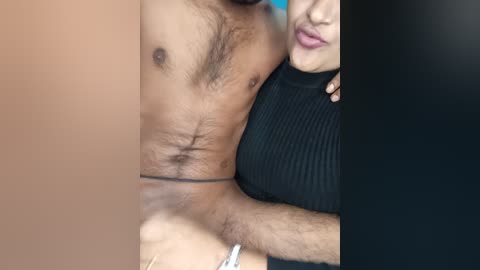 Video showing a close-up of a topless man with a hairy chest, wearing only a thin black string, hugging a woman with a black sweater, both with light skin tones.