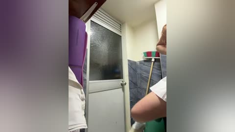 A video of a person wearing a white shirt and jeans, cleaning a bathroom door with a sponge. The bathroom has a purple wall, white door, and a green shower curtain.
