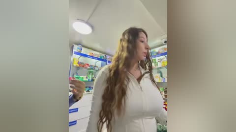 Video of a woman with long, wavy brown hair in a white jacket, standing in a brightly lit, neatly organized pharmacy, shelves filled with colorful products.