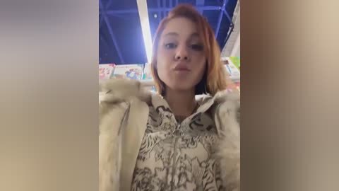 Video of a young woman with straight, shoulder-length red hair, wearing a light gray, patterned blouse, standing in a brightly lit, colorful bookstore with shelves of books in the background.