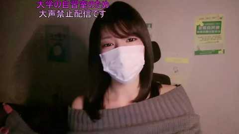 A video of a young Asian woman with long black hair, wearing a white face mask, gray off-the-shoulder sweater, and a black top, sitting indoors with posters in the background.