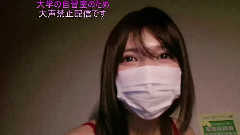 A video of an East Asian woman with long black hair, wearing a white surgical mask and a red tank top, against a plain background. Text in Japanese is overlaid in pink.
