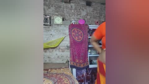 Video of a woman in a vibrant orange dress, standing in a rustic room with a patterned purple drape, yellow cloth, and a brick wall background.