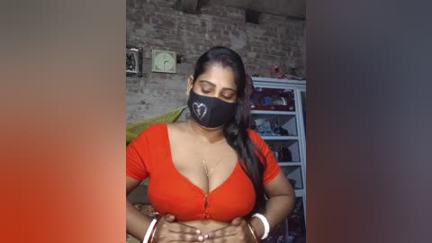 Video of a dark-skinned woman with long black hair, wearing a black face mask, red crop top, and bracelets, standing in a rustic room with a brick wall, clock, and white shelves.