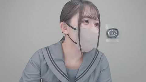 Video of a young Asian woman with straight, dark hair, wearing a gray school uniform with a white mask, standing against a plain, light-colored wall with a clock.