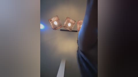 Video of a dimly lit interior with a rectangular, geometric chandelier hanging from a high ceiling. The background is soft, blurred, and grey. The foreground shows a partially visible, muscular male figure with a dark complexion, wearing a dark shirt, standing in the frame.