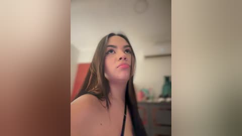 Video of a young woman with light brown skin and long brown hair, wearing a black top, looking contemplative in a dimly lit bathroom with a red door and a green plant.