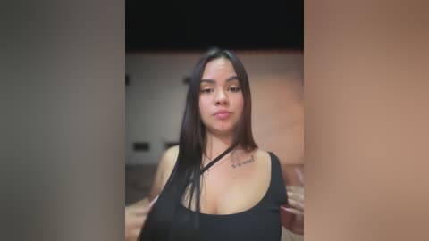 Video of a Latina woman with long black hair, medium skin tone, and a black tank top, standing in a dimly lit room with beige walls and a blurred background.