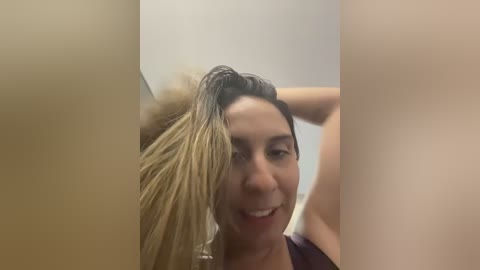 Video of a smiling woman with wet, blonde hair, wearing a dark shirt, in a bathroom with beige walls and a mirror.