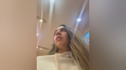 A video of a young woman with long, straight blonde hair, wearing a white sweater, captured from a low angle, emphasizing her face. The background shows a modern, well-lit room with recessed ceiling lights.