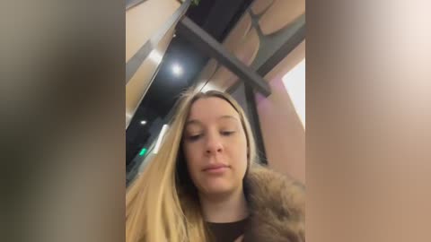 Video of a young blonde woman with fair skin, wearing a brown coat, seated in a dimly lit, modern train car with beige walls, overhead luggage racks, and a green light in the background.