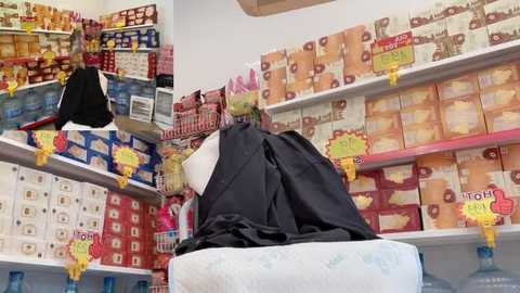 Video of a supermarket aisle filled with colorful packaged foods, including pasta, cereals, and snacks. A person in a black apron and pink hairnet is working behind a white counter.