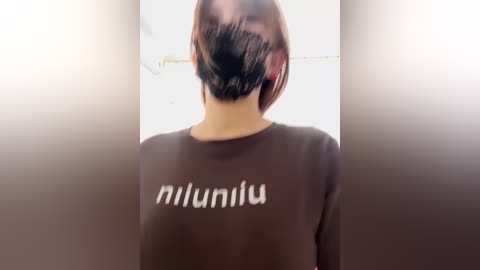 Video of a person with a medium build, wearing a black face mask and a brown \"niluminus\" t-shirt. Background is blurred, with a white wall and ceiling.