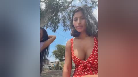 Video of a Latina woman with medium-dark skin and shoulder-length black hair, wearing a low-cut red dress with white polka dots, standing outdoors under a tree with a bright blue sky.