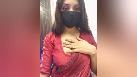 Video of a South Asian woman with medium skin tone, wearing a red saree, black face mask, and purple eyeshadow. She covers her chest with her hand, revealing a wedding ring. Background is blurred with warm tones.